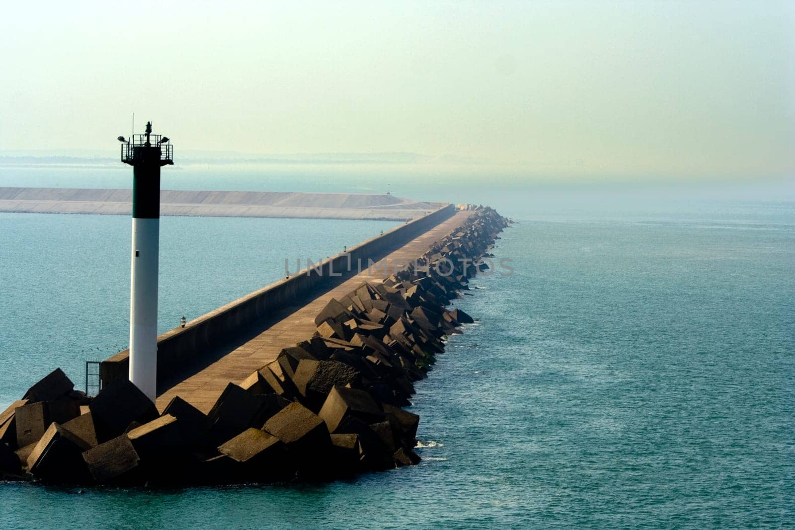 Lighthouse structure at the Port of Dunkirk (France) at the end of a road extending into the sea with sharp-edged rocks breaking the waves. Haze and sharp contrast create a striking image