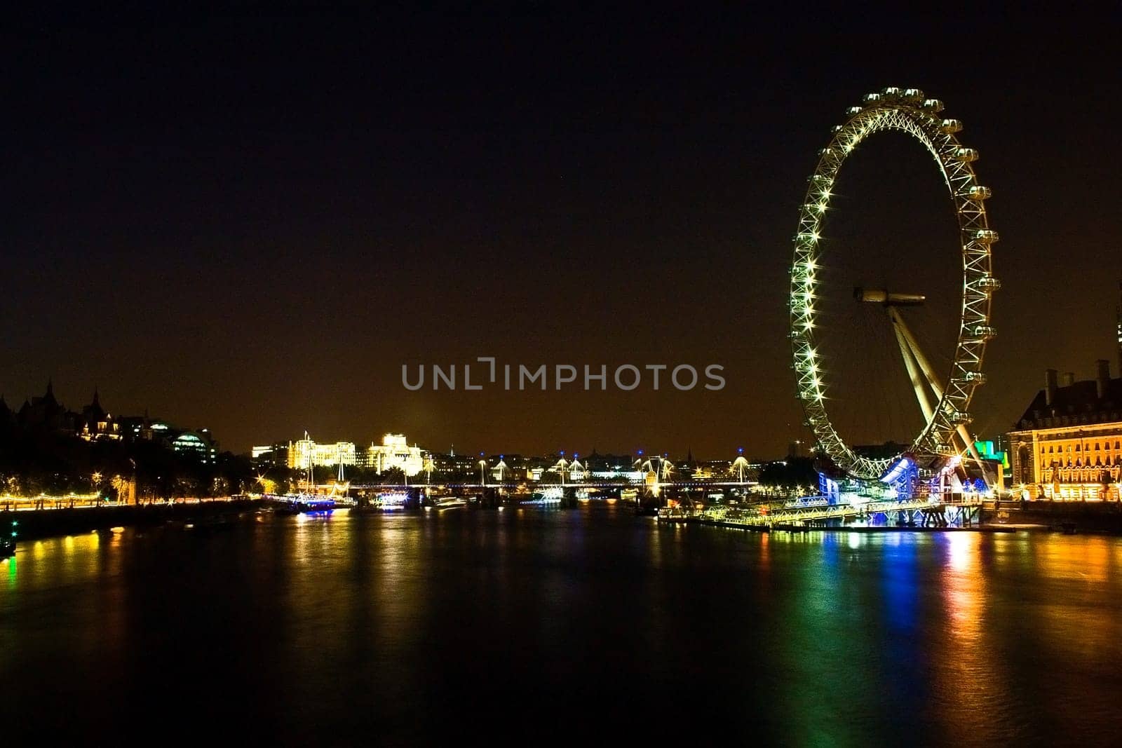 The London Eye (Millenium Wheel) lights up the London city skyline with green, blue and orange reflections in the Thames river water