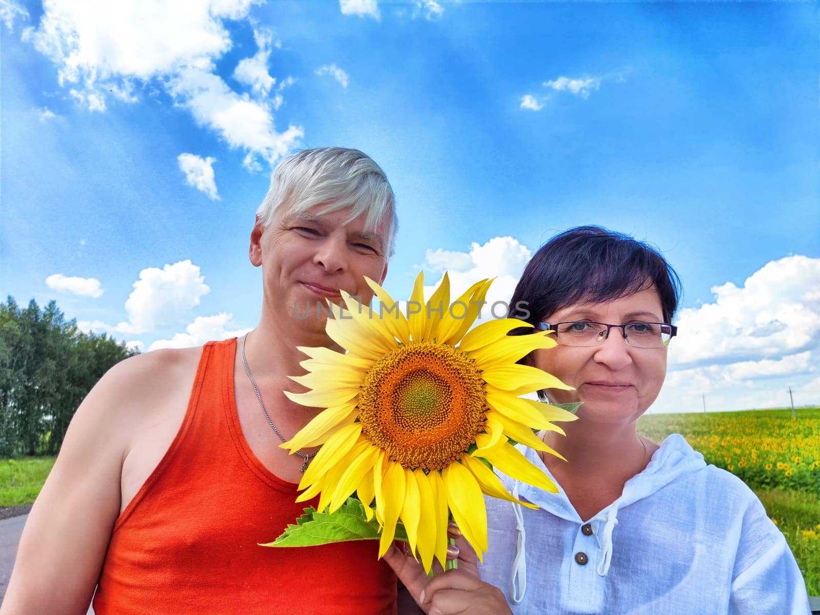 Middle-aged couple smiles together holding a sunflower, enjoying their travels in a beautiful outdoor setting with vibrant greenery and a clear blue sky.
