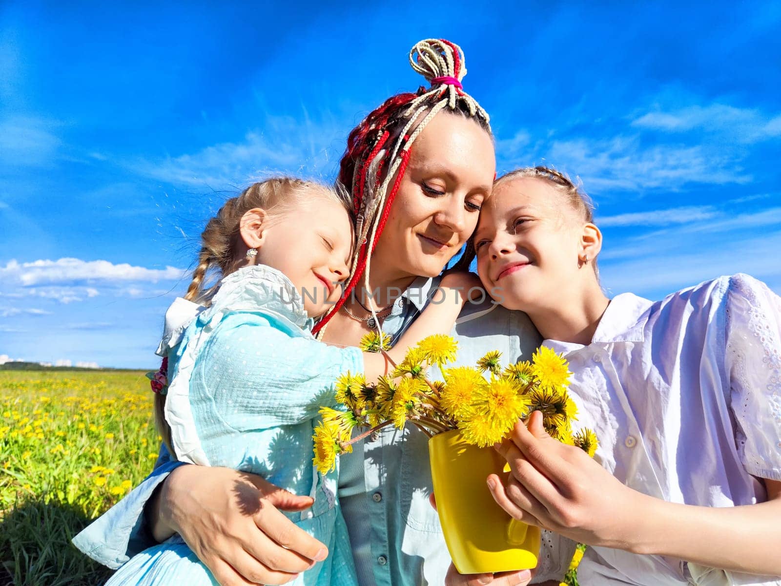 In a bright, sunny field filled with dandelions, a mother shares a joyful moment with her two daughters as they pick flowers.