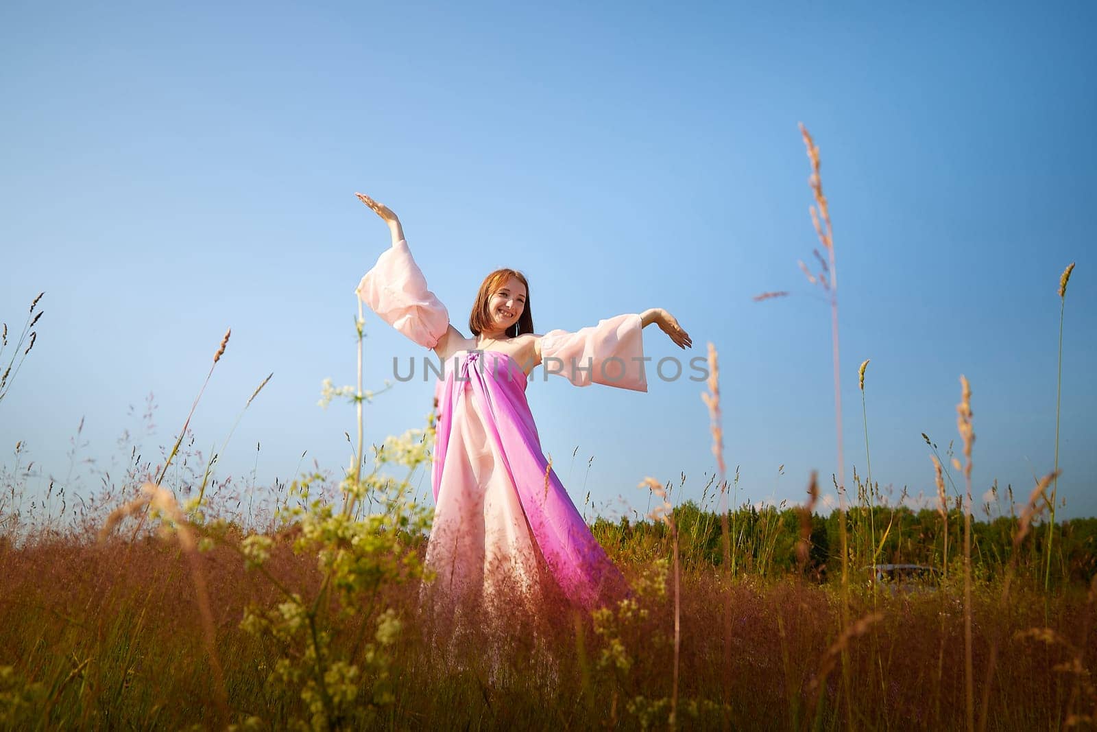 A joyful woman wearing a colorful dress dances gracefully in a sunlit meadow, surrounded by wild grasses under a clear blue sky.