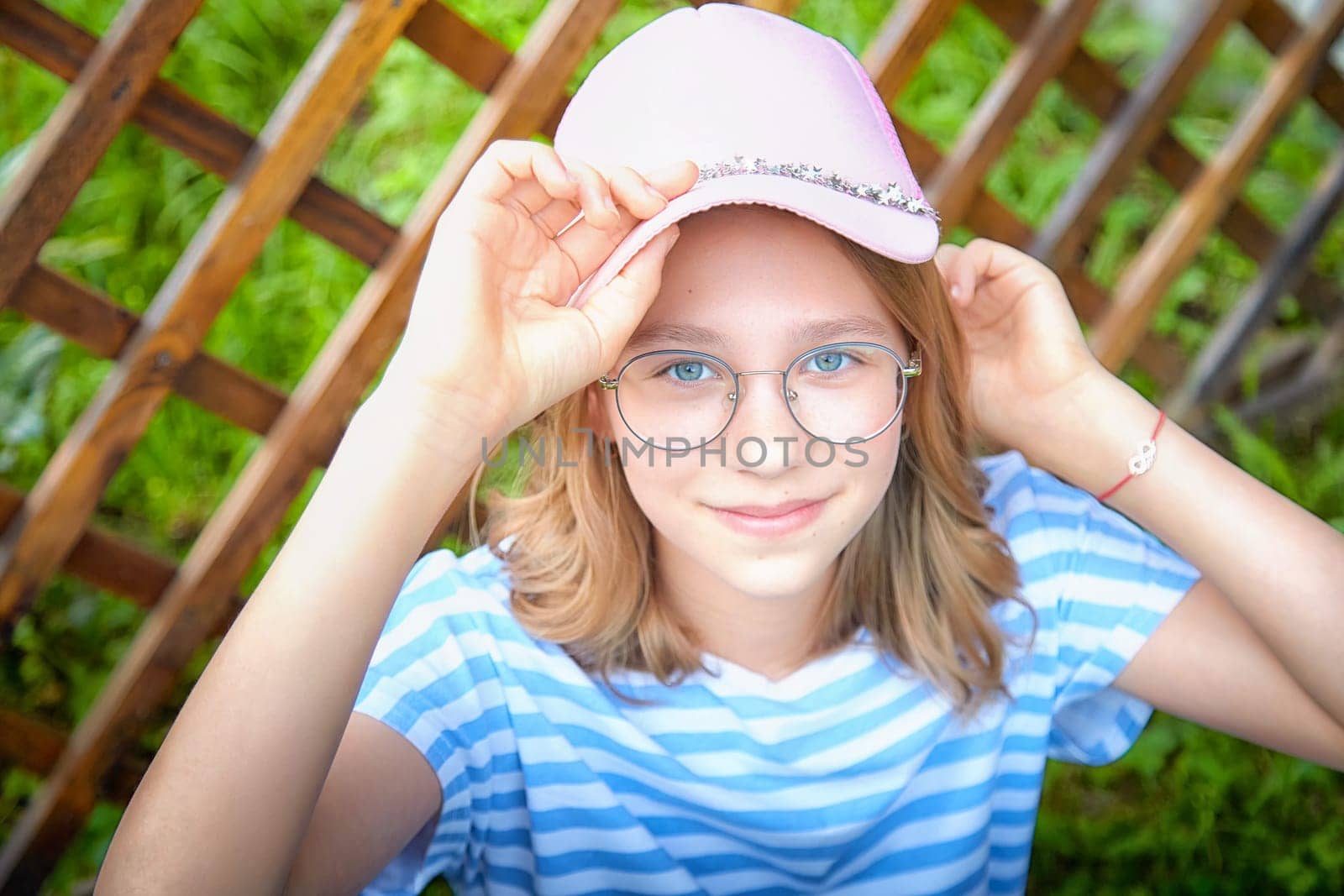 A cheerful girl with glasses and a pink cap playfully adjusts her hat, surrounded by lush greenery in a bright garden.