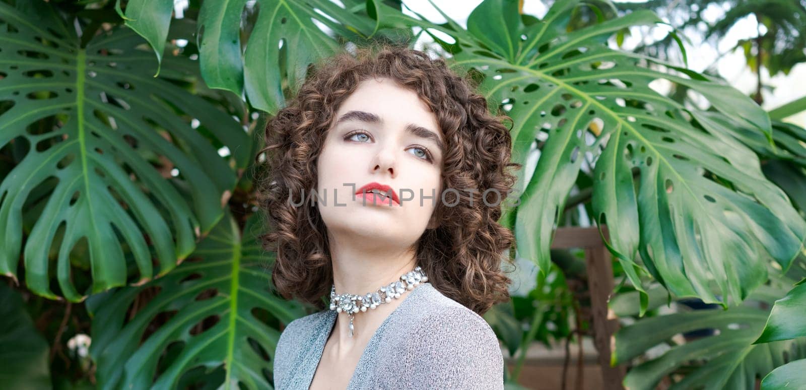 Woman with curly hair is wearing a necklace and a red lipstick. She is standing in front of a large green plant