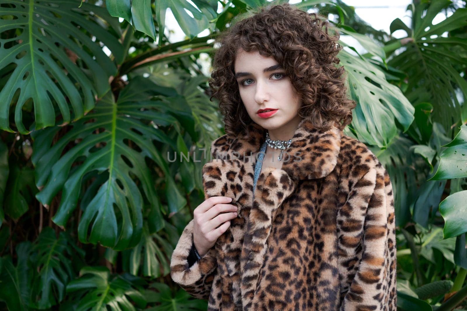 Woman wearing a leopard print coat and a necklace. She is standing in front of a green plant