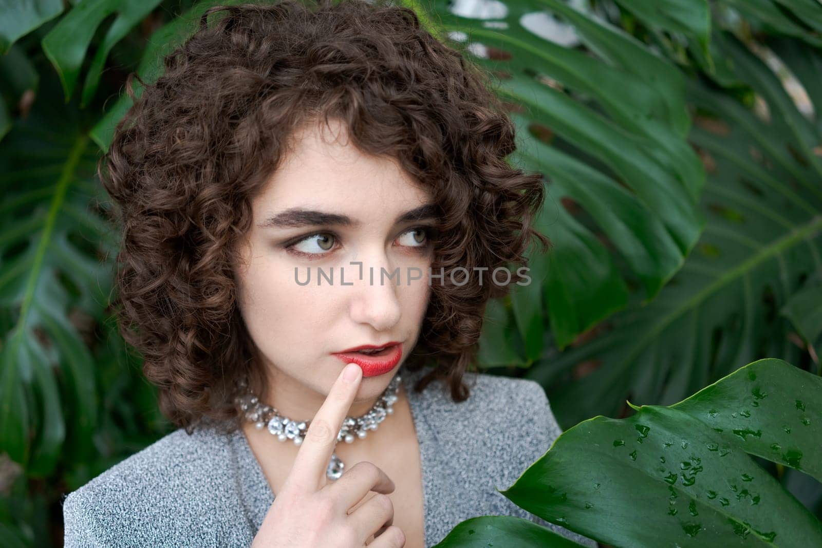 Woman with curly hair and a necklace is looking at the camera. She is wearing a gray jacket and a red necklace