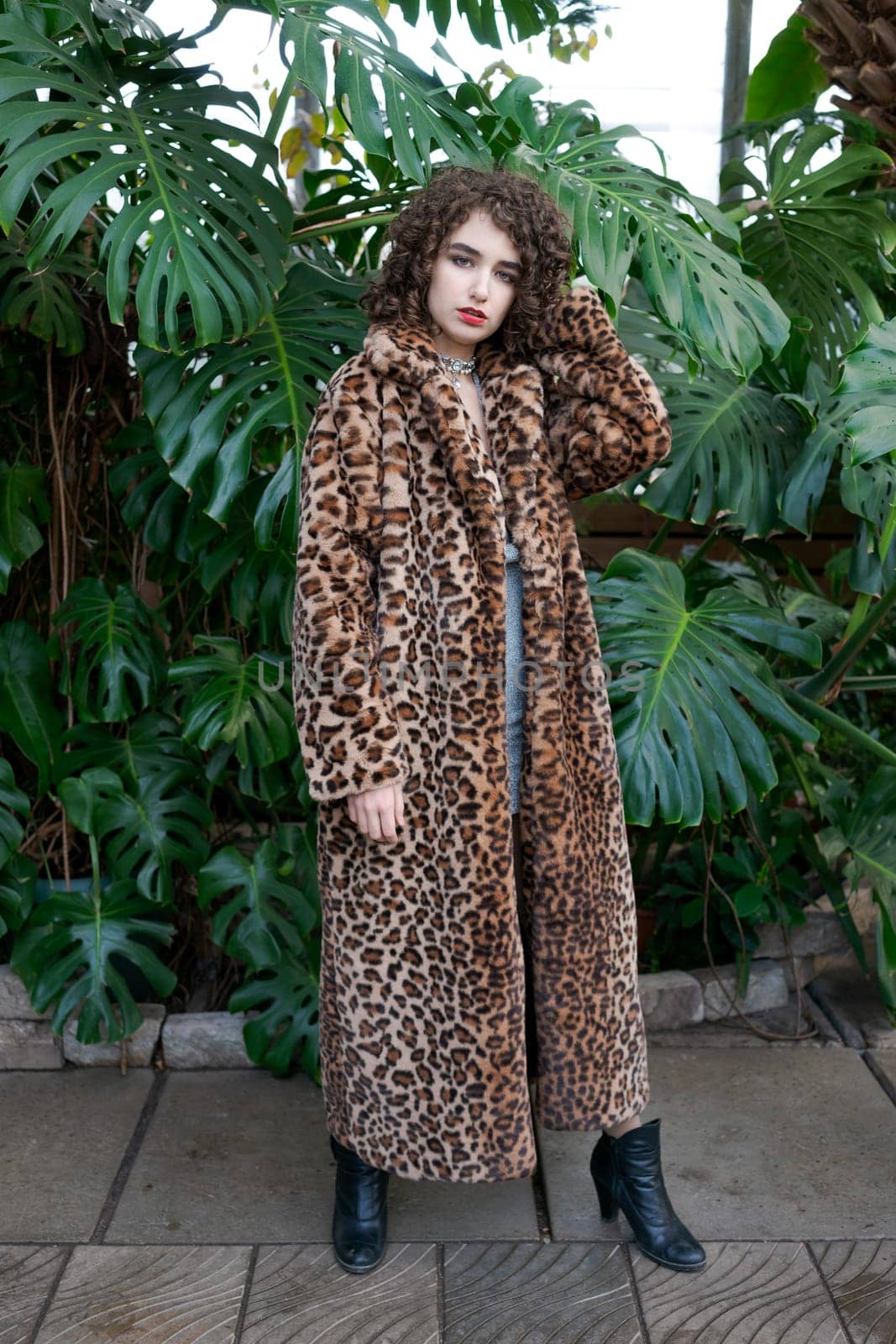 Woman wearing a leopard print coat poses in front of a green plant. She is wearing black boots and a necklace