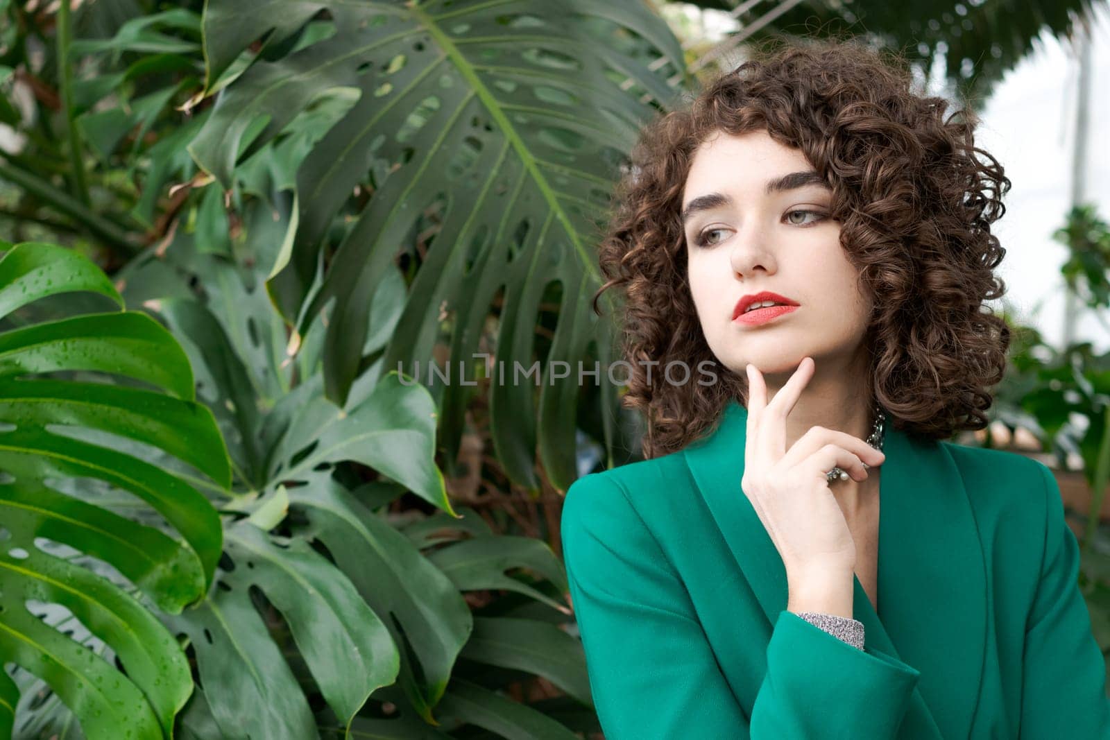 Woman with curly hair is wearing a green jacket and looking at the camera. She is wearing a necklace and a red lipstick