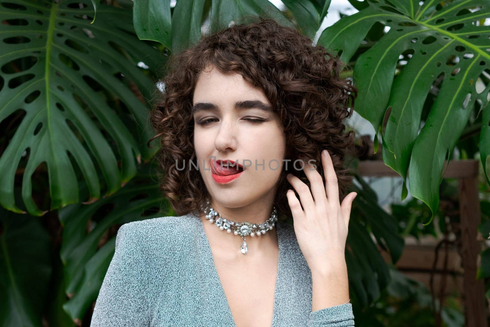 Woman with curly hair is wearing a necklace and a blue dress. She is licking her tongue