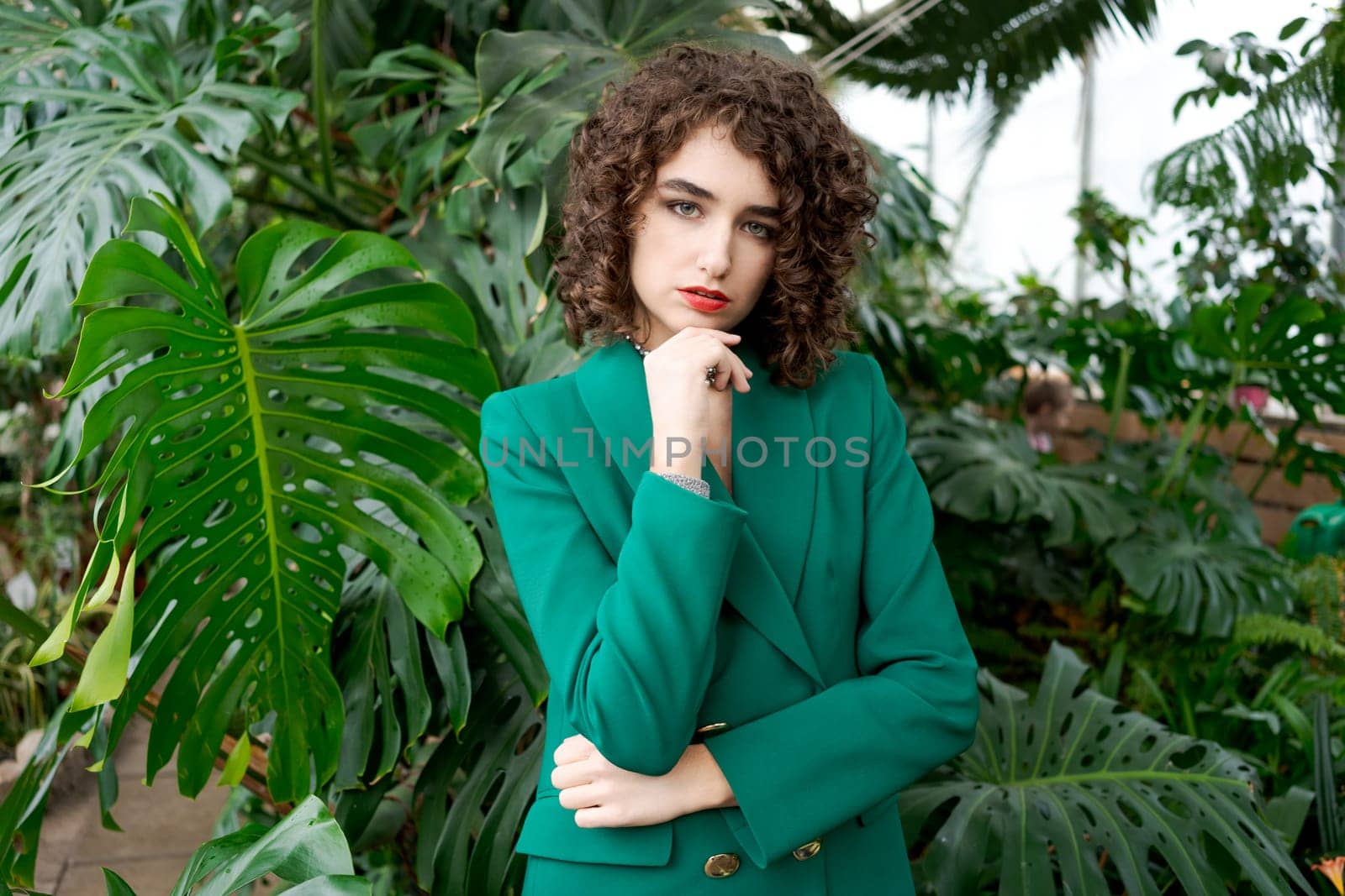 Woman in a green jacket is standing in front of a plant. She is wearing a red lipstick
