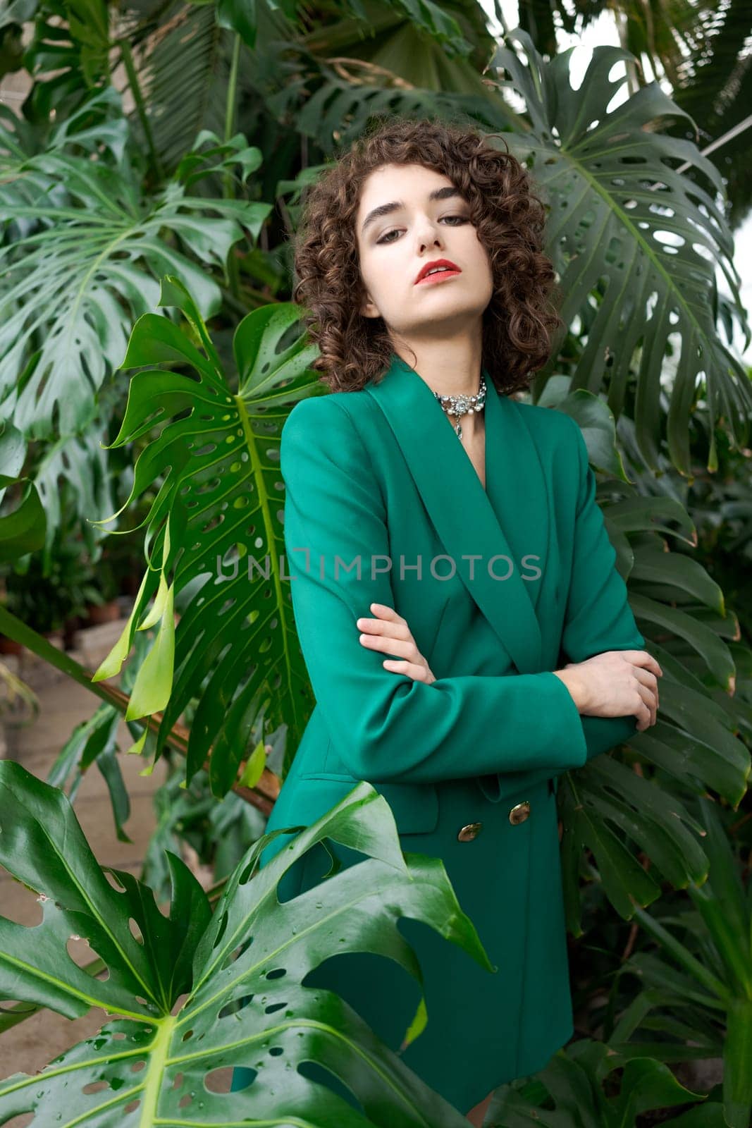 Woman in a green dress is standing in front of a plant. She is wearing a necklace and a red lipstick