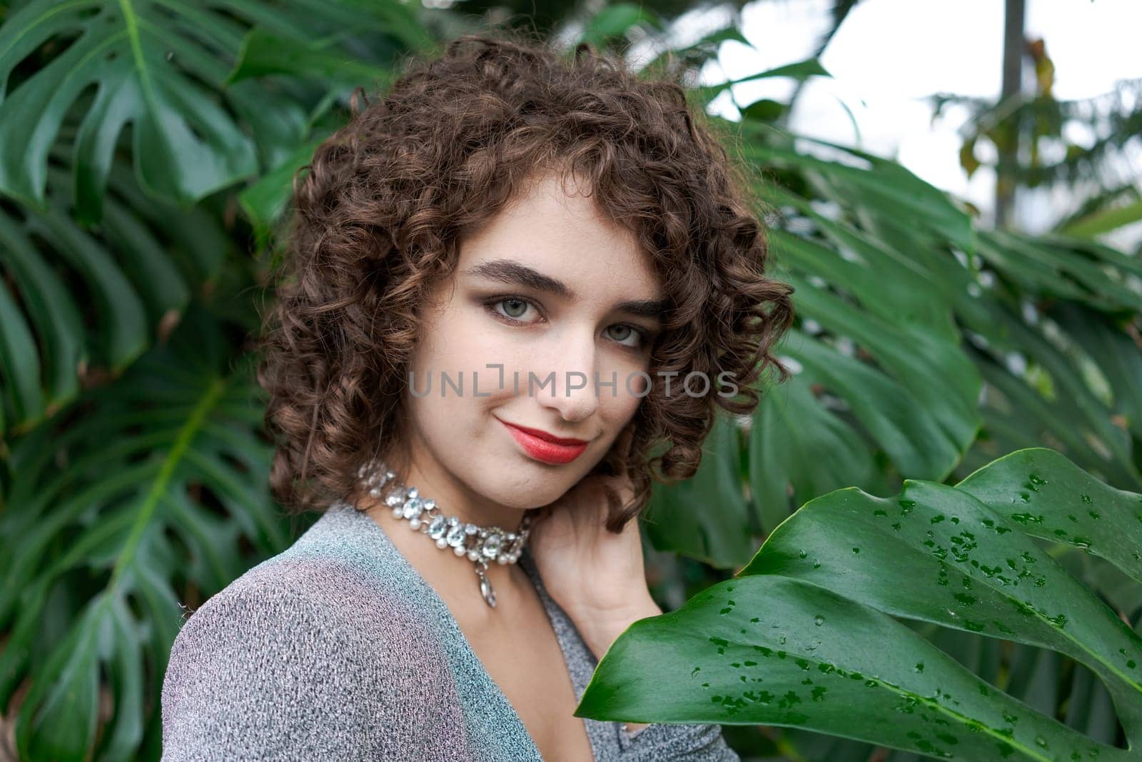 Woman with curly hair is wearing a necklace and a red lipstick. She is standing in front of a green plant
