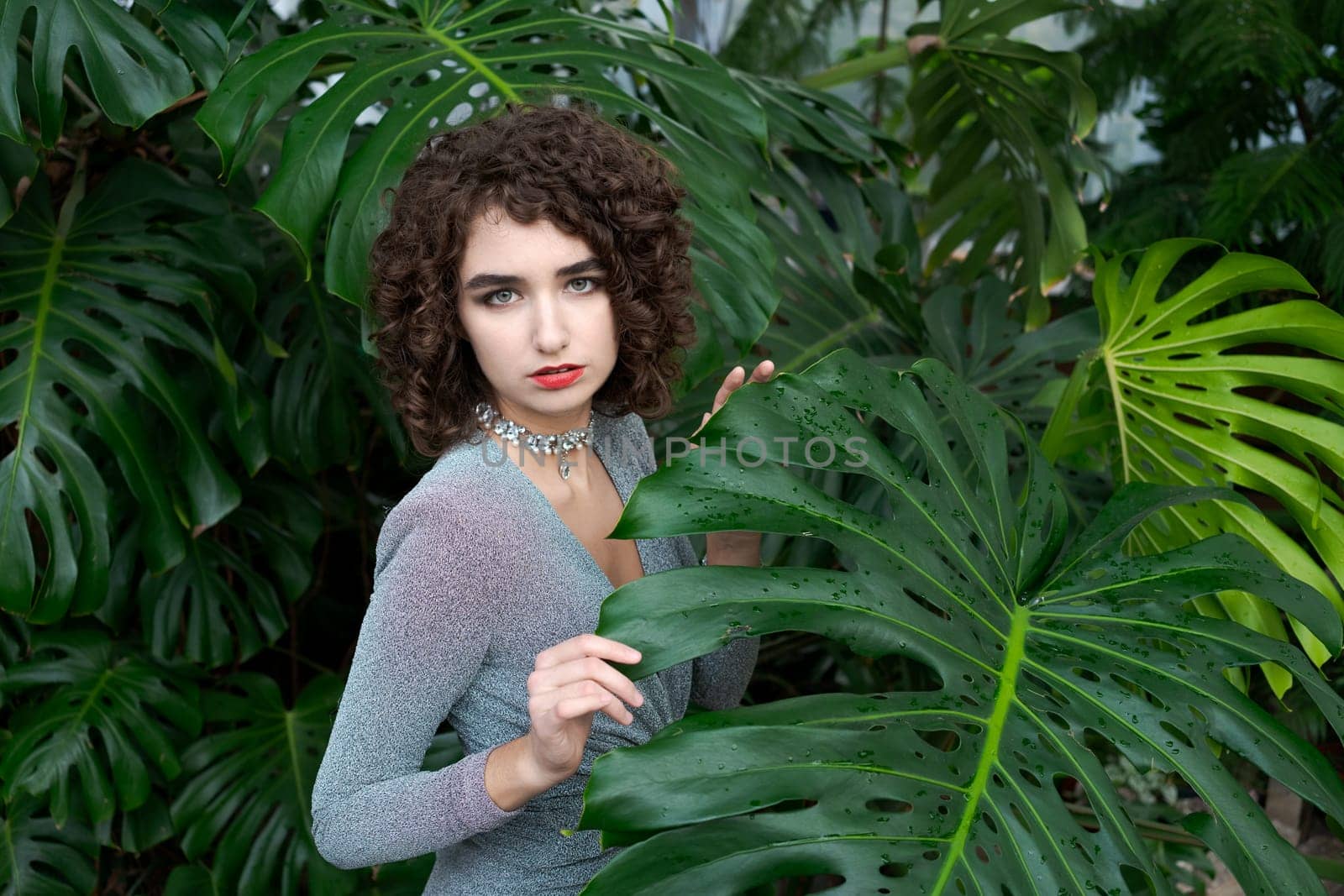 Woman is standing in front of a large leafy plant. She is wearing a silver necklace and a red lipstick