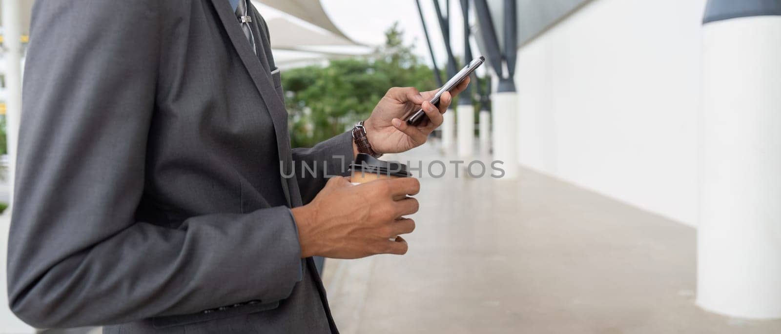 A businessman in a suit holding a coffee cup and checking his smartphone while standing outdoors in a modern setting, combining work and refreshment.