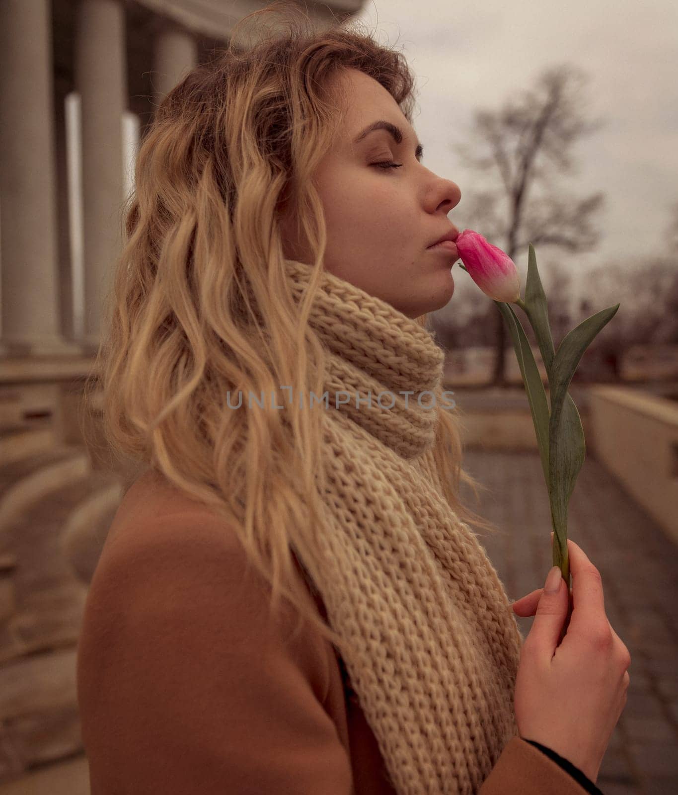 close-up of a blonde with a flower near her lips. portrait of a girl, near her face she holds one pink tulip. she stands against the background of an 18th century colonnade. dressed in a beige coat
