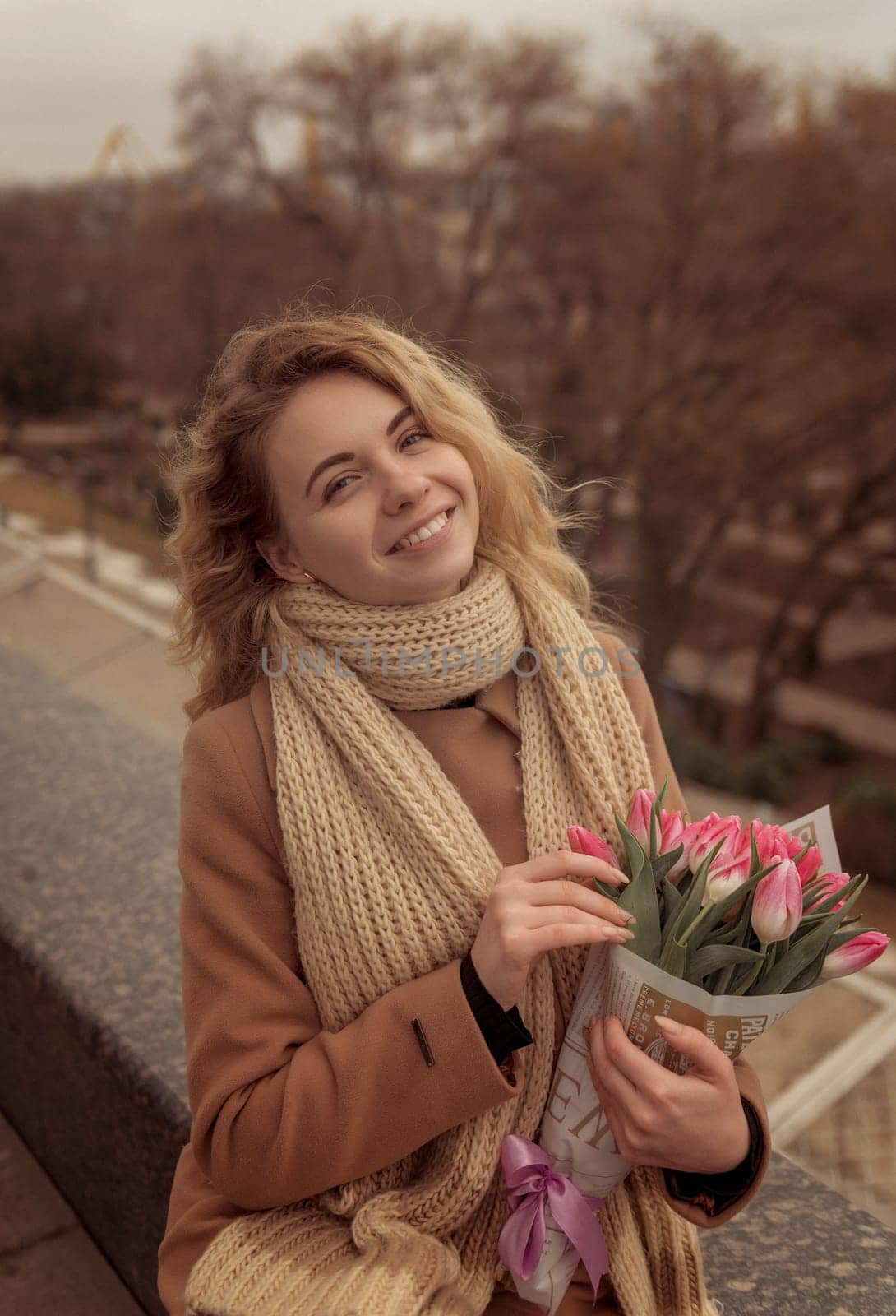 Close-up of a blonde holding a bouquet of pink tulips. The bouquet is wrapped in craft paper. She is laughing and looking at the camera. Her hair is flying. She is wearing a beige classic coat and a warm scarf.