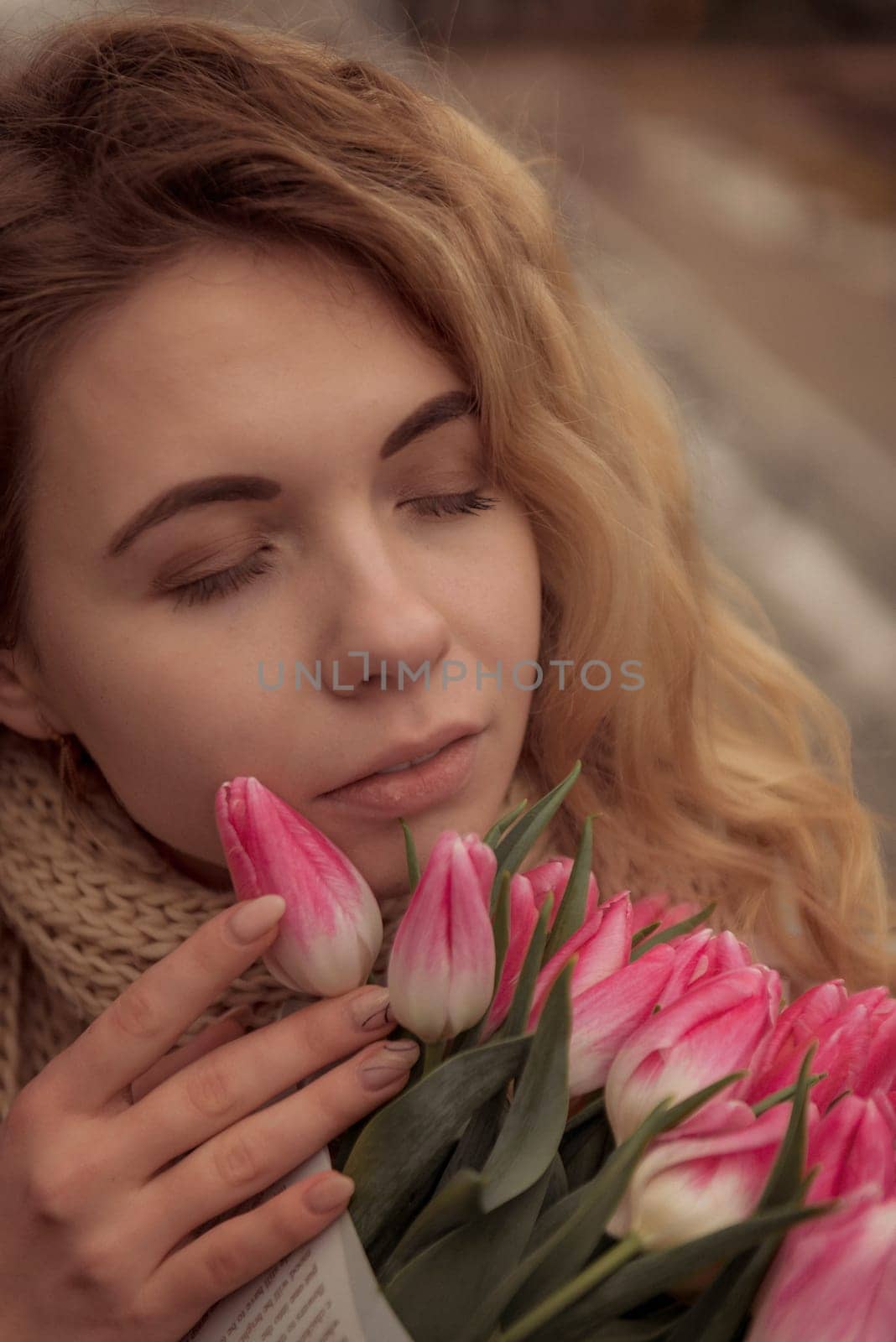 Close-up of blonde with bouquet of pink tulips. bouquet is wrapped in craft paper. she is dreaming her eyes are closed. her hair is flying. dressed in beige classic coat, warm scarf.
