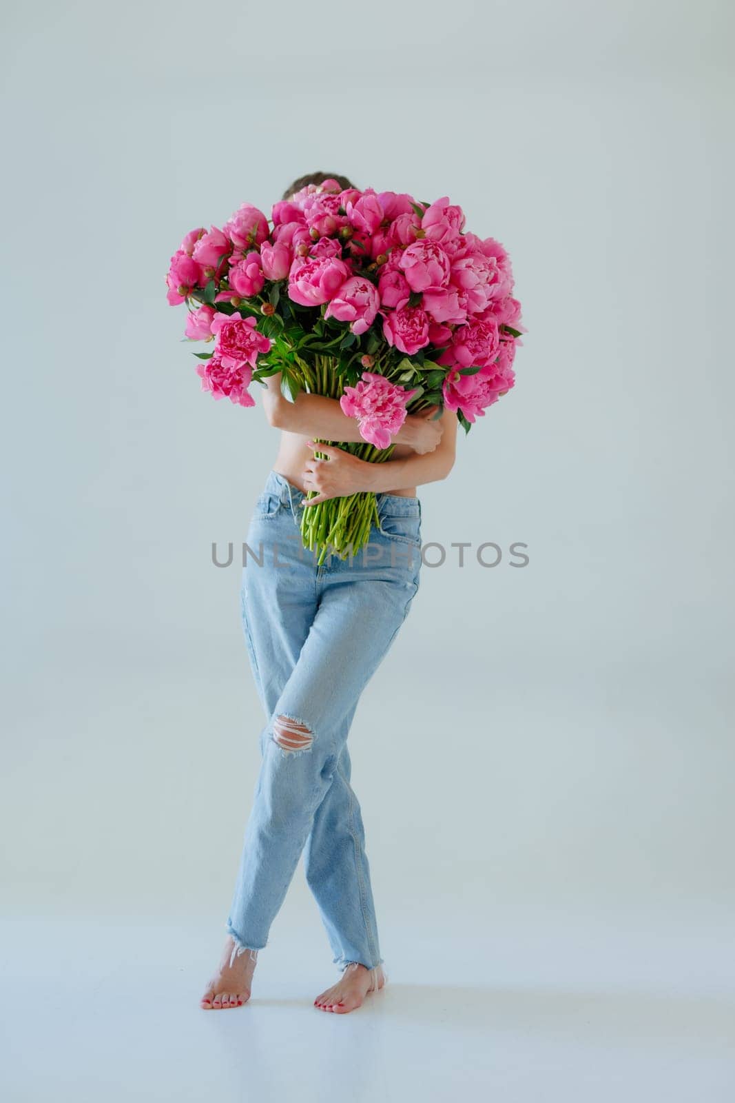 A woman in jeans holds a huge bouquet of pink peonies, completely hiding her upper body. The minimalist background, barefoot pose, and bright flowers create a stylish, artistic, and modern aesthetic.