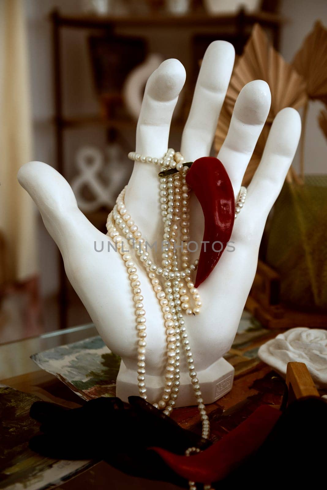 plaster hand holding pearl necklace and red hot pepper. close-up hand made of white plaster, paint brushes nearby. concept hot classic, white and red