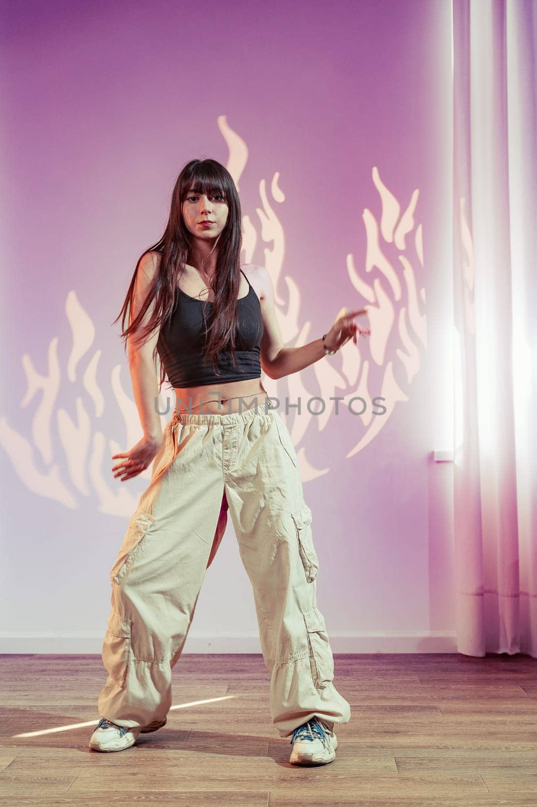 A woman performs an expressive dance routine in a spacious studio. She wears loose beige pants and a black top, moving confidently under soft, colorful lights that enhance the atmosphere.