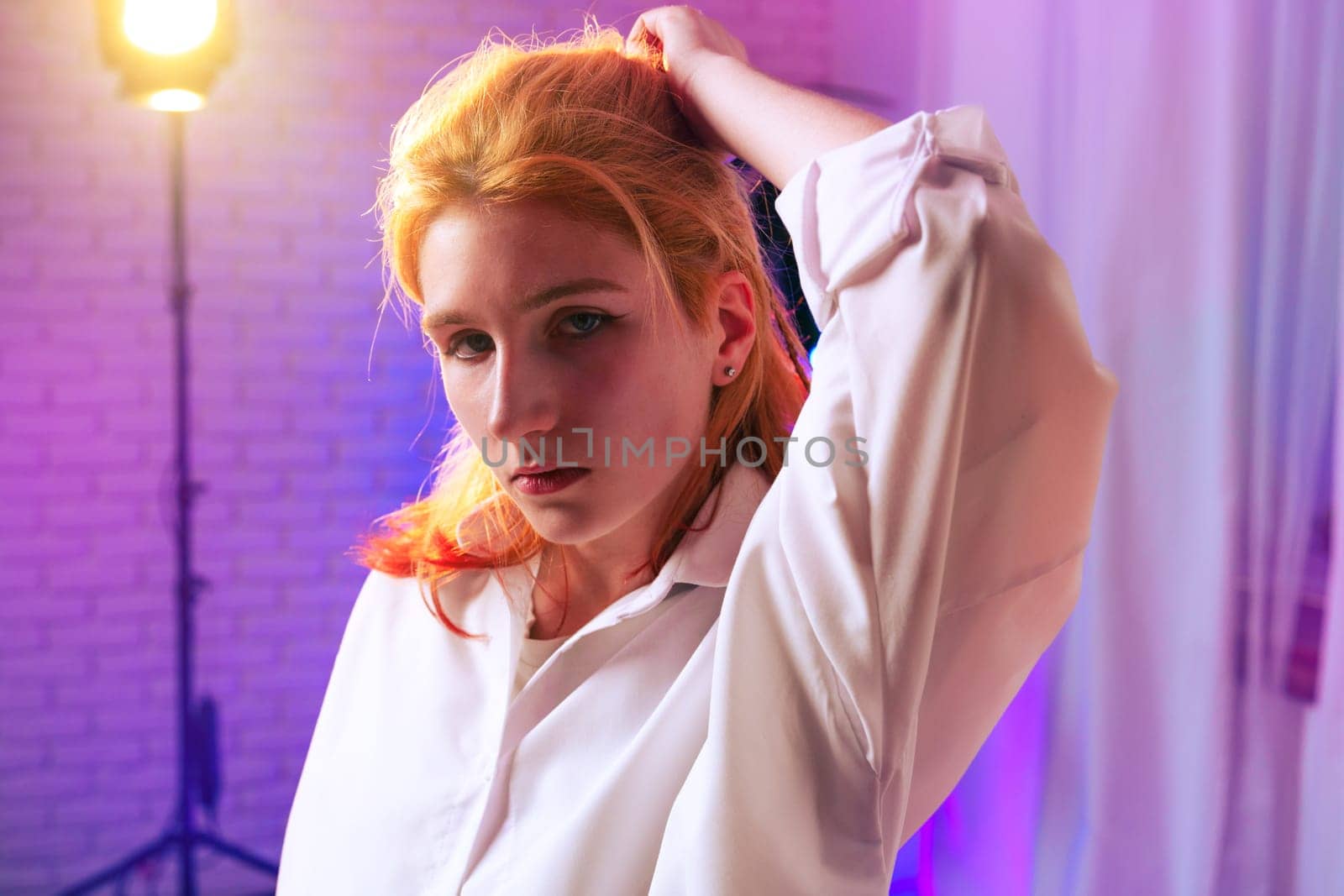 A young woman stands confidently in a modern indoor space, wearing a stylish oversized shirt. Colorful lighting highlights her striking features and creates an artistic atmosphere.