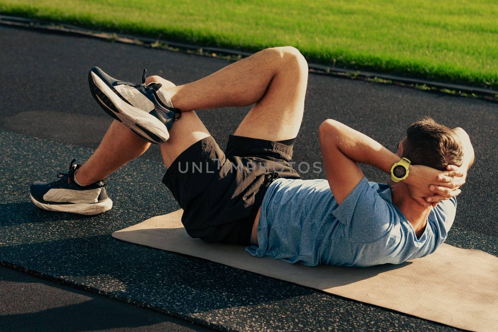 A man performs abdominal exercises on a mat at a track field in the morning. His focus is on enhancing his core strength while enjoying the warm weather and open space.