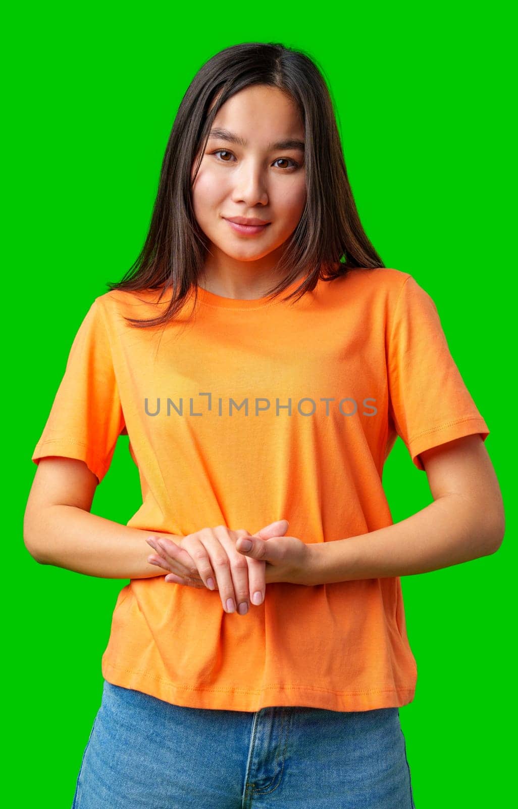 A young woman stands confidently with her hands clasped together, wearing an orange t-shirt. The bright green background highlights her friendly demeanor and the vivid color of her outfit.