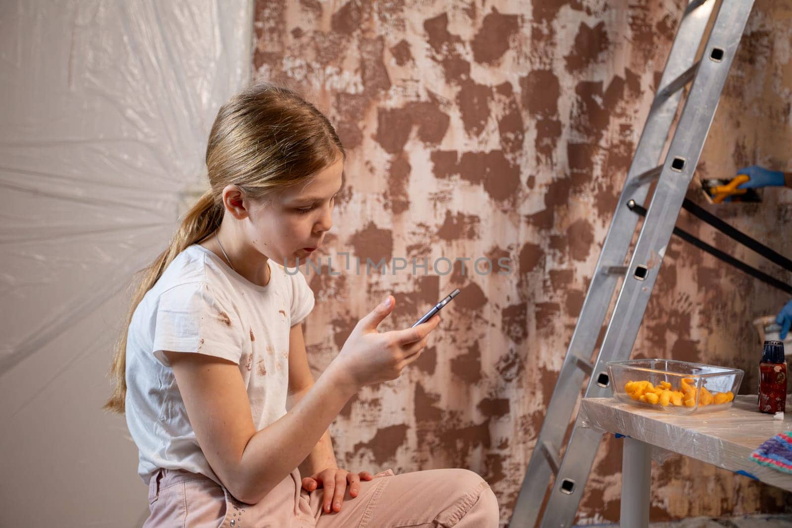 A focused young girl sits in a creative setting, engaging with her phone and surrounded by art supplies.