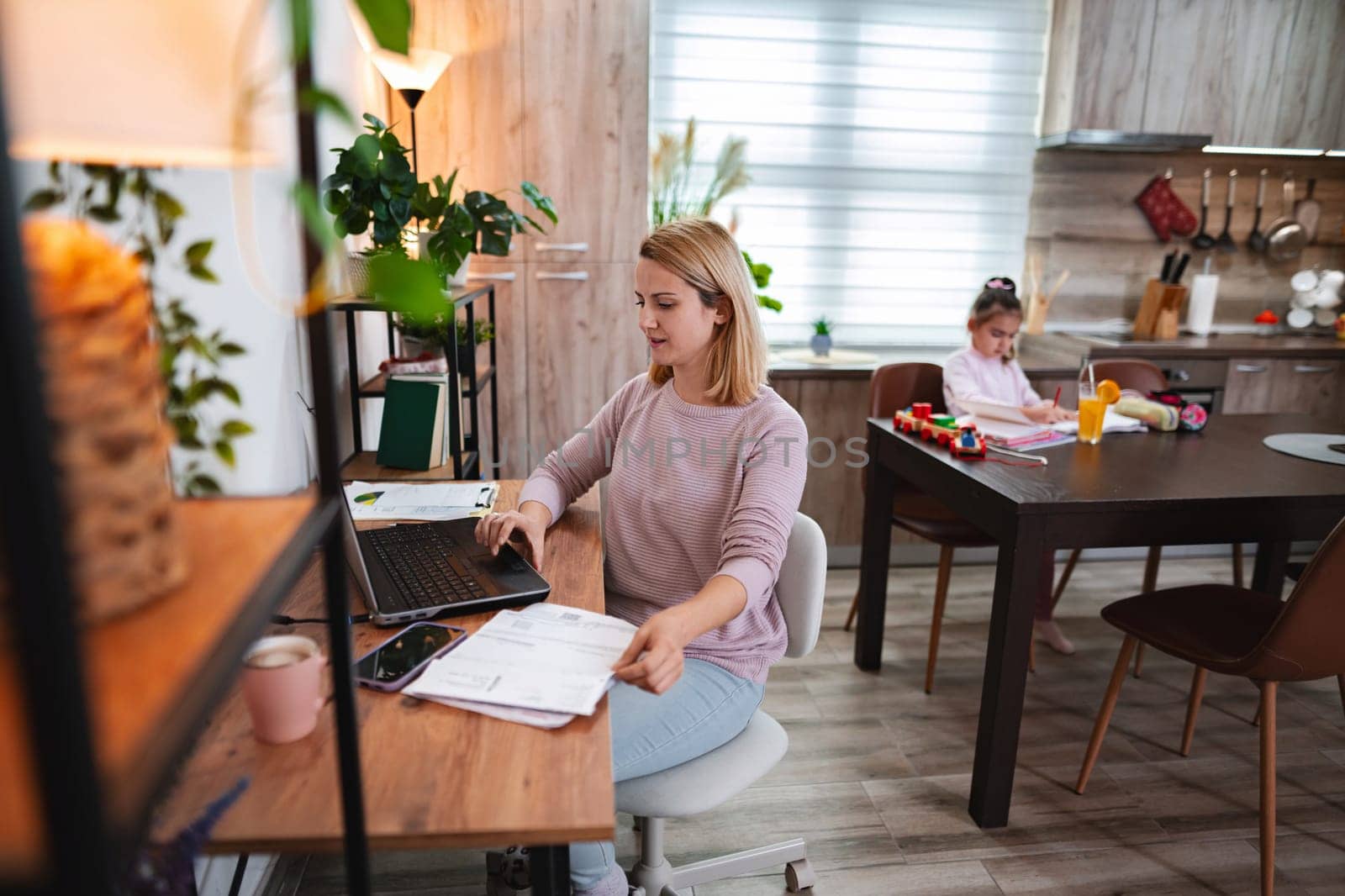 A woman works on her laptop as a child plays in a warm kitchen.