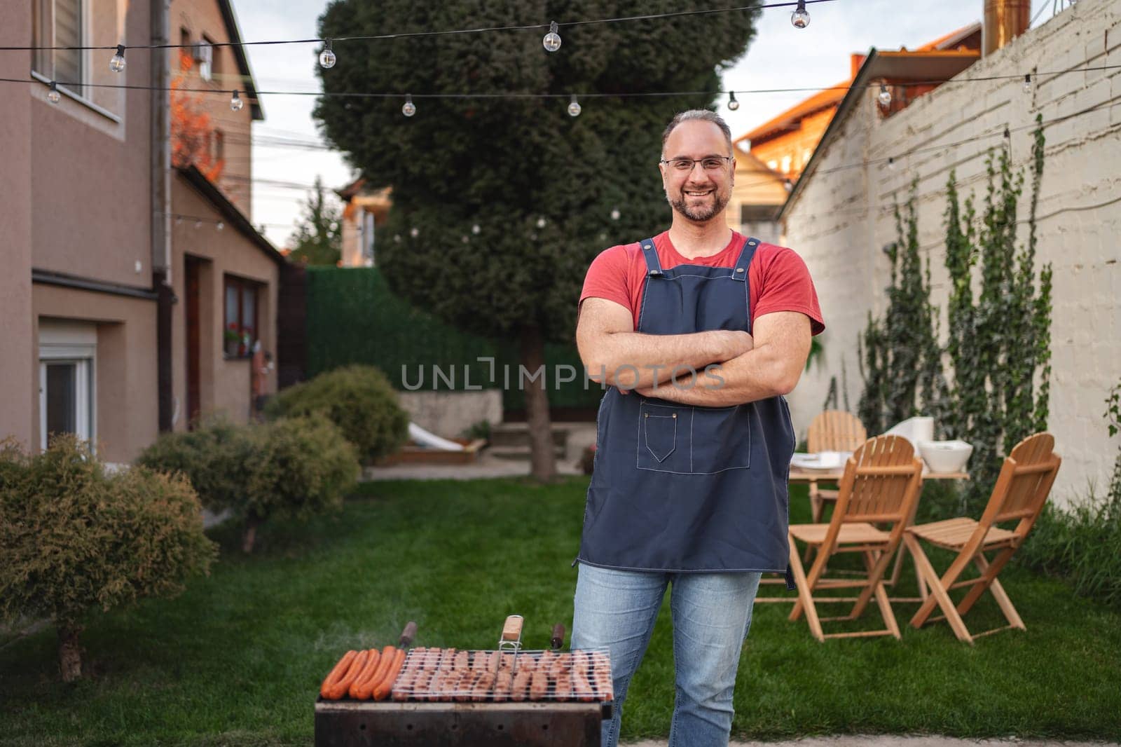 Grill master stands proudly in a lush backyard while cooking delicious sausages.