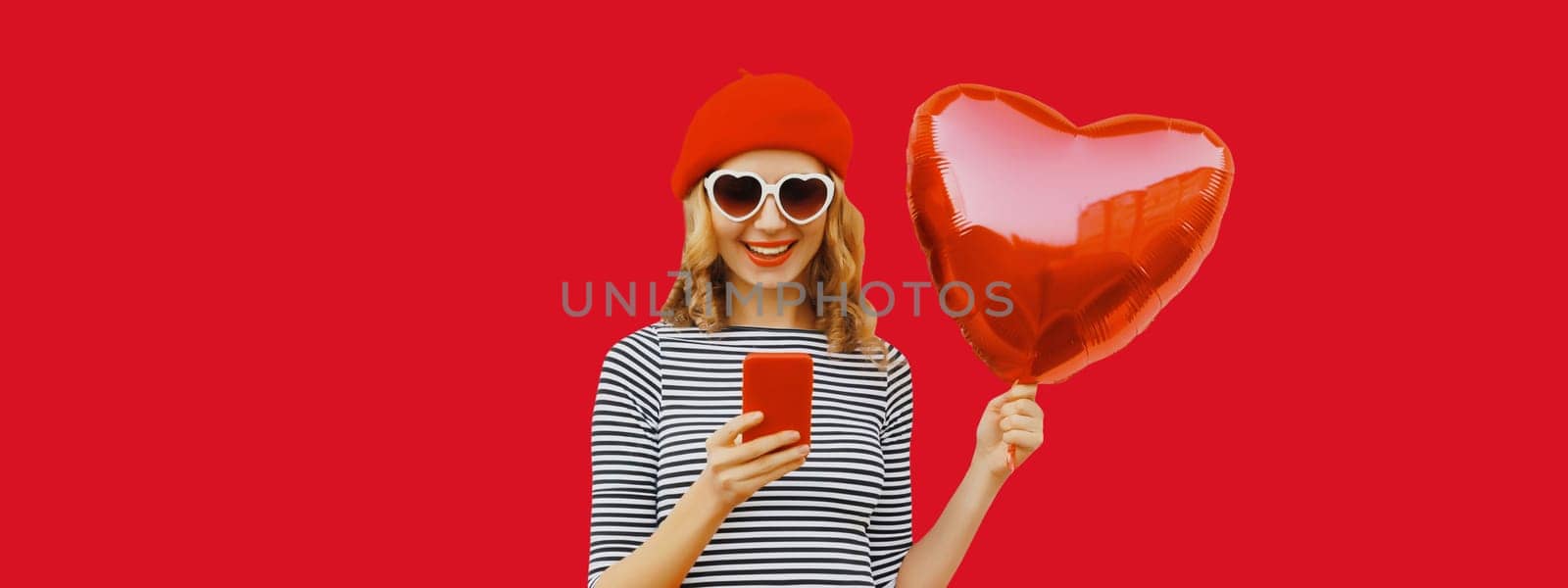 Portrait of beautiful happy smiling young woman with smartphone holding red heart shaped balloon wearing french beret, sunglasses on studio background