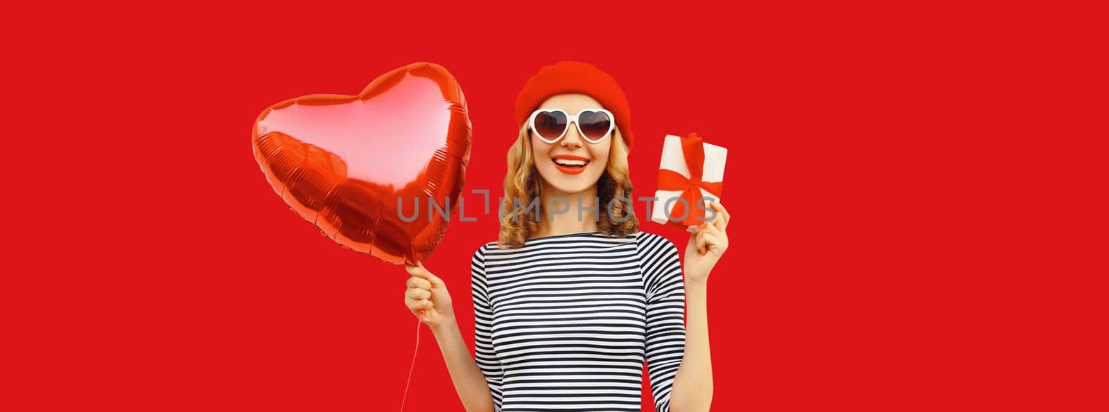 Portrait of happy laughing young woman holding red heart shaped balloon and gift box wearing french beret hat isolated on studio background