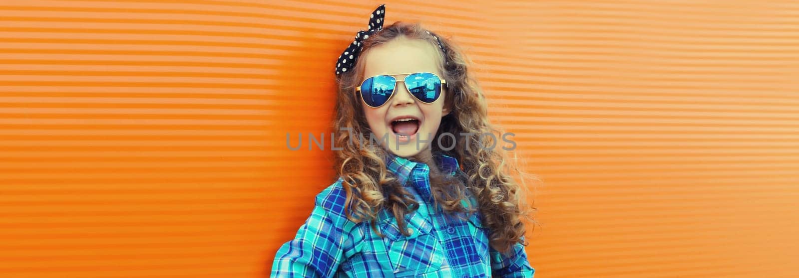Happy cheerful kid girl with curly hair, joyful little child having fun on city street on orange wall background