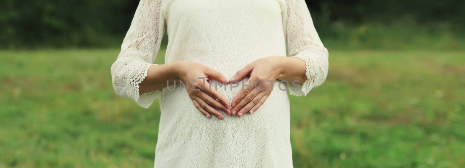 Pregnancy, maternity, close-up happy pregnant woman makes heart sign on her belly with her hands in summer park