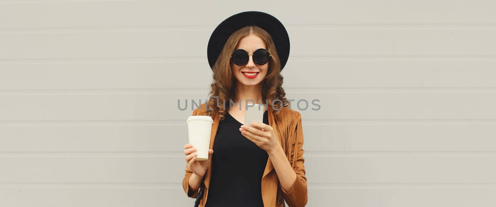 Stylish happy smiling young woman with phone, beautiful girl holding smartphone, cup of coffee wearing black hat standing on city street on gray background