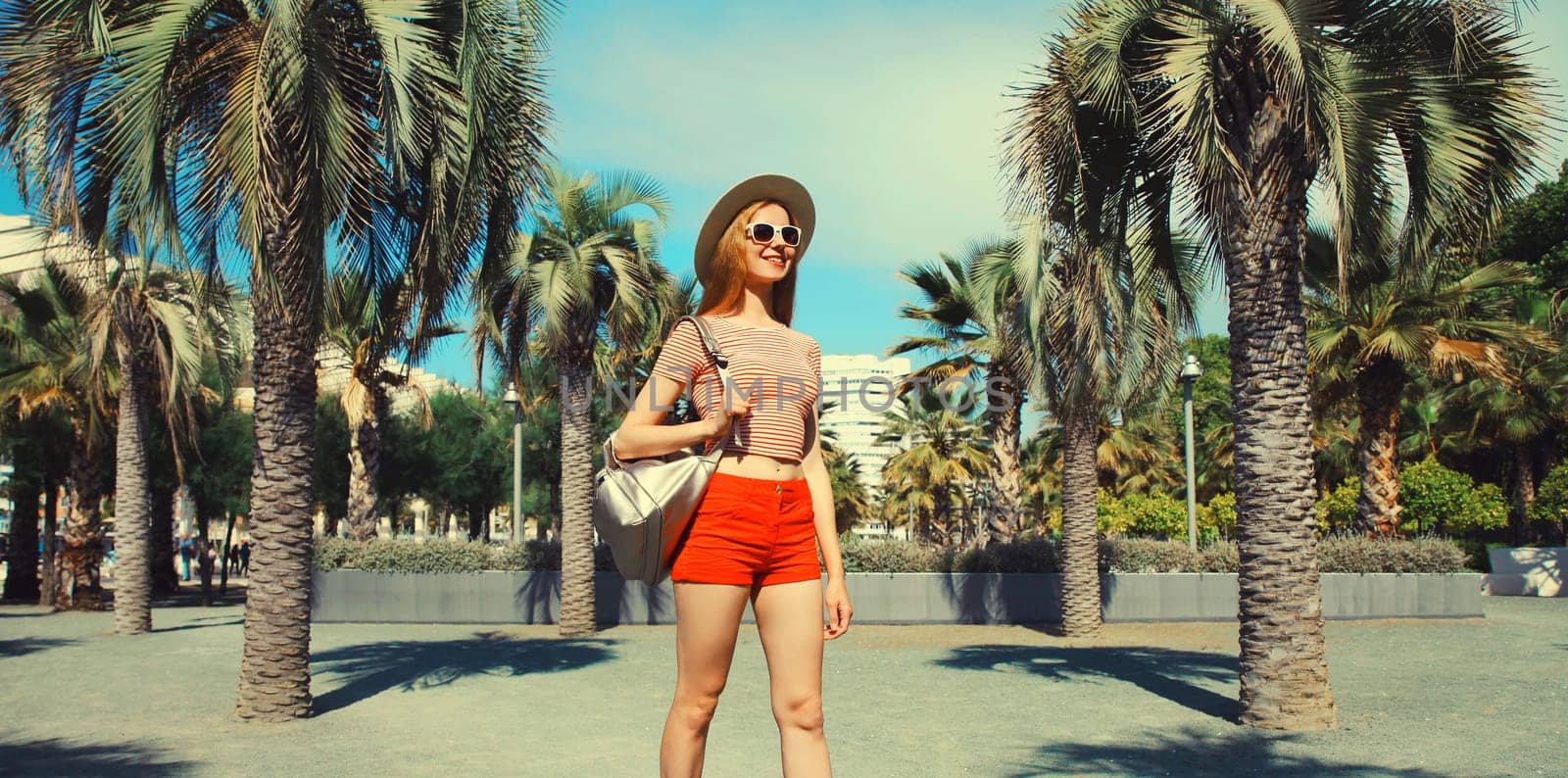 Beautiful happy young woman in summer park wearing a backpack, tourist hat against a palm trees background