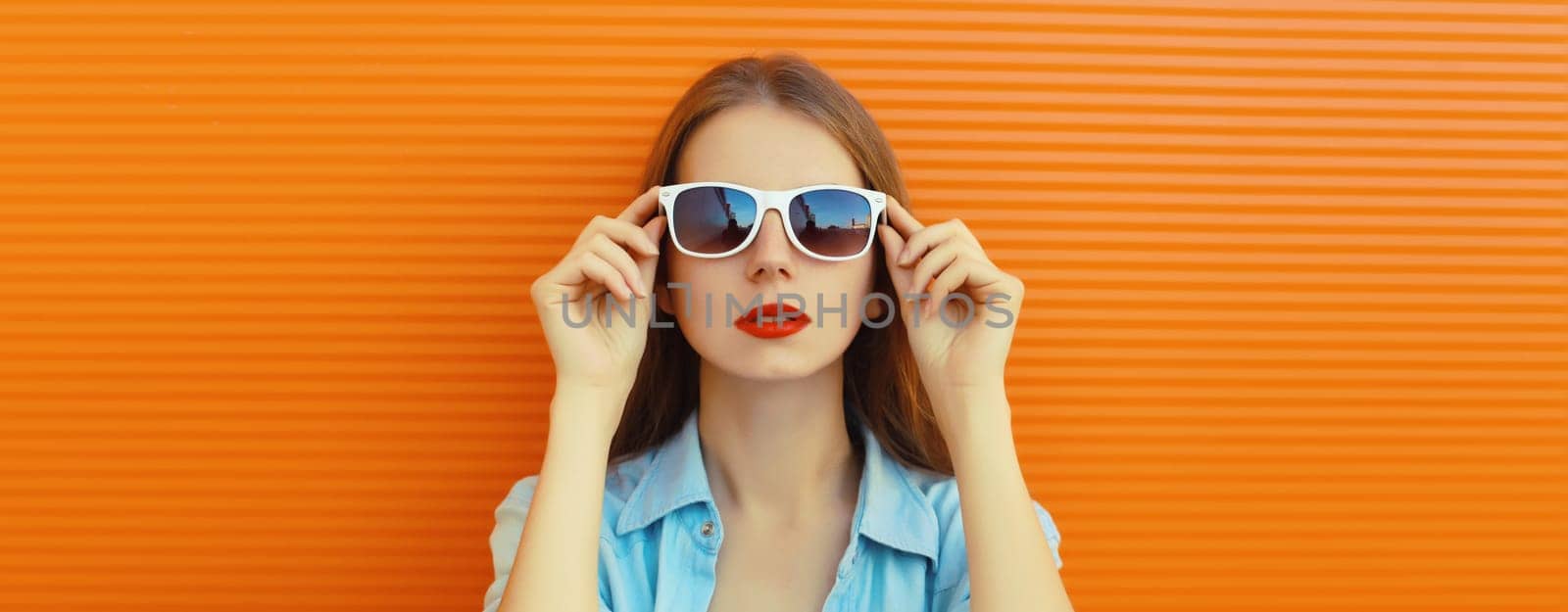 Close-up portrait of modern stylish young woman in white glasses, casual clothing posing on vivid orange wall background