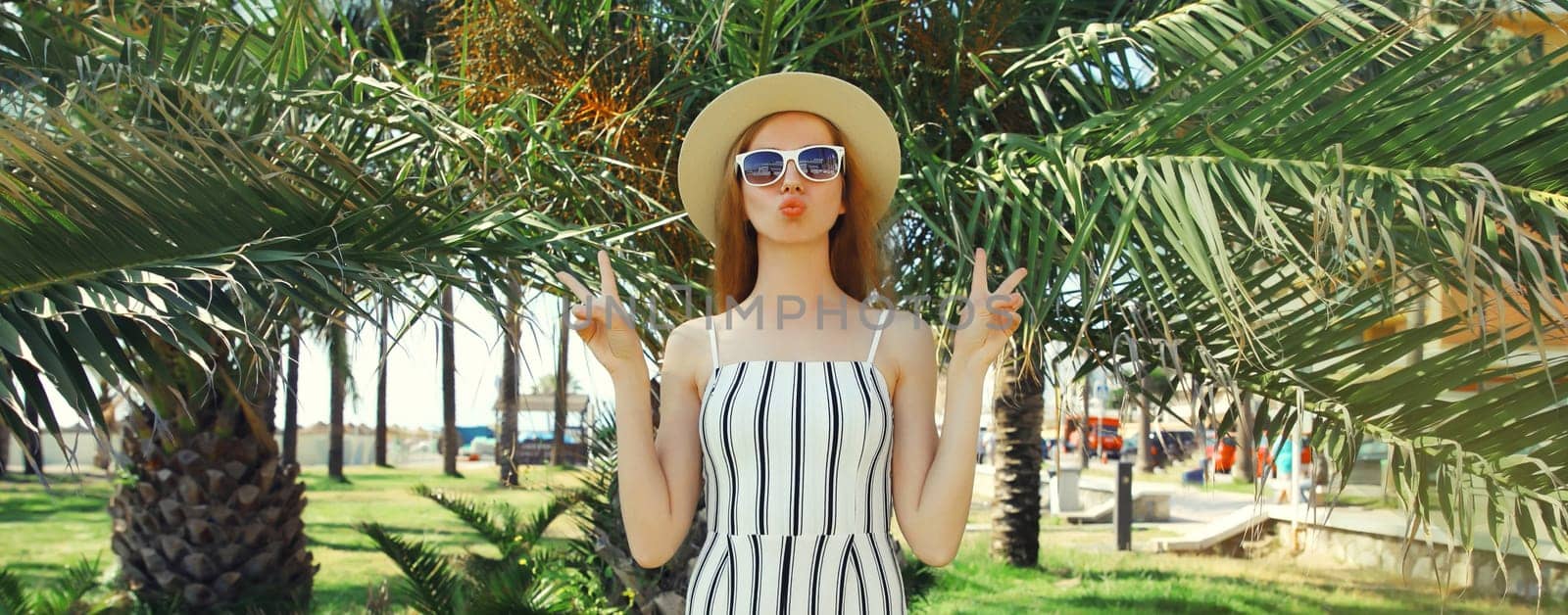 Summer outfit, stylish beautiful young woman posing in white striped jumpsuit standing in green park with palm trees background