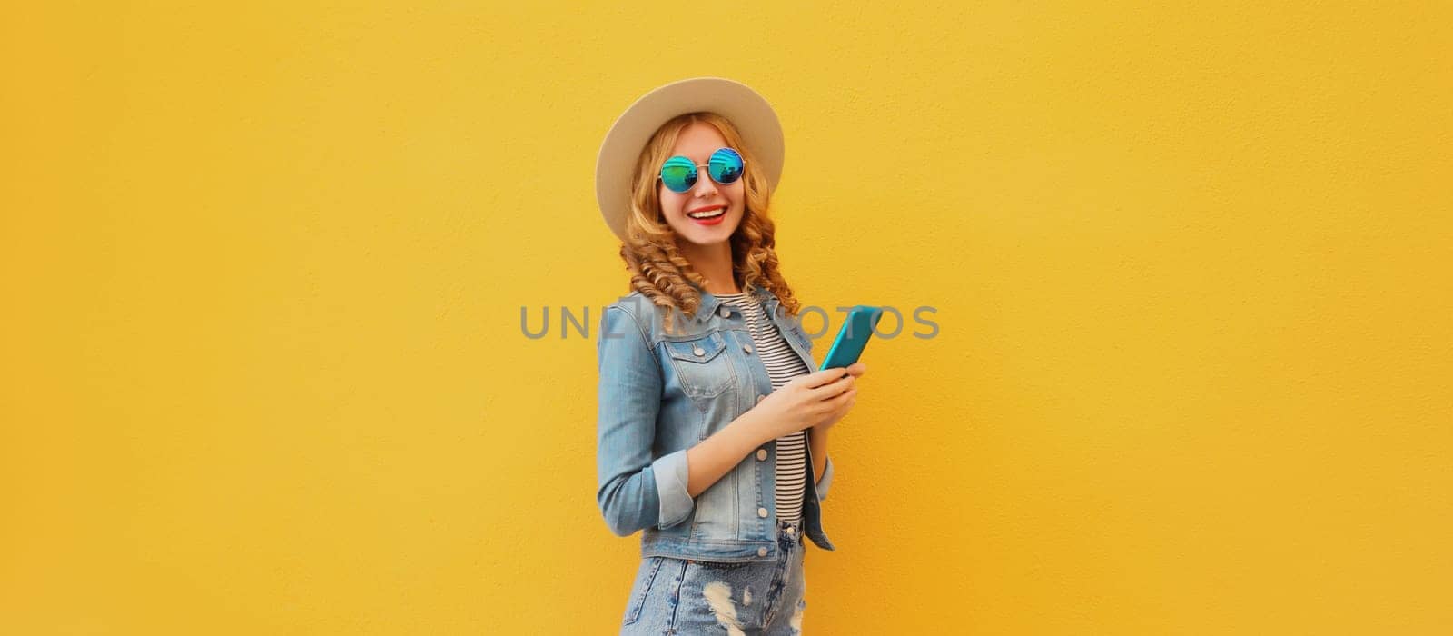 Summer modern happy young woman holding a smartphone wearing a straw tourist hat, denim clothing posing on yellow wall background