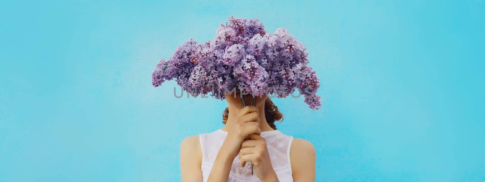 Close-up of woman covering her head with spring bouquet of fresh lilac flowers on blue background, nature, bloom