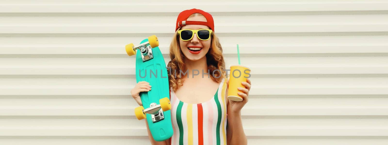Summer portrait of happy cheerful stylish young woman with skateboard and cup of fresh juice in colorful clothes standing on white background