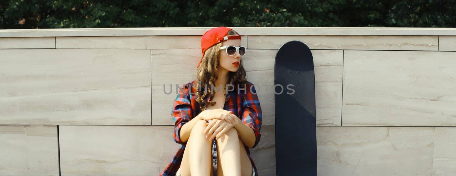 Stylish young woman sitting with skateboard in casual clothes looking away, modern teenage girl posing in the city