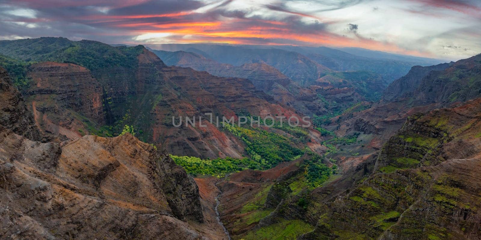 Aerial view of Waimea Canyon's red and brown cliffs, green vegetation, and deep valleys under a vibrant sunset with orange, pink, and purple clouds.