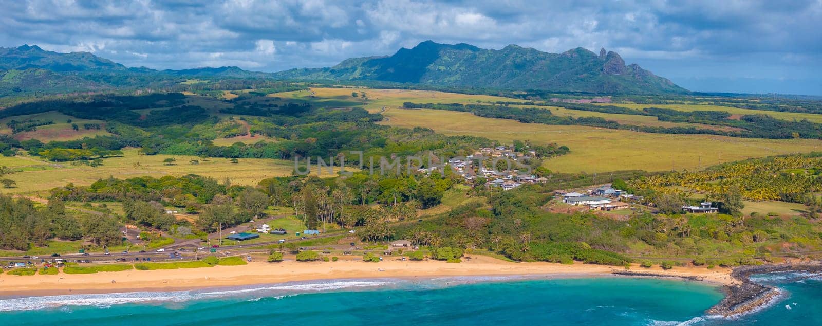 Aerial view of Kauai Island, Hawaii, showing a golden sandy beach, turquoise waters, a coastal community, lush greenery, and the Napali cliffs.