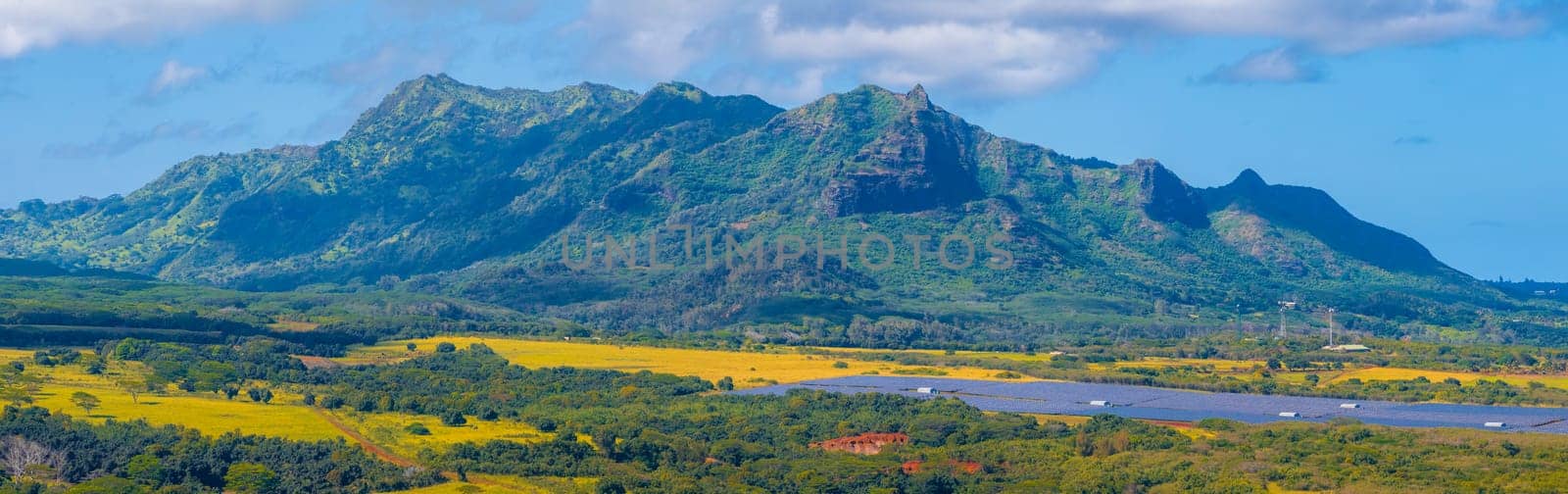 Aerial view of Kauai Island, Hawaii, showing green mountains, rugged ridges, fields with mixed vegetation, and a solar farm under a partly cloudy sky.