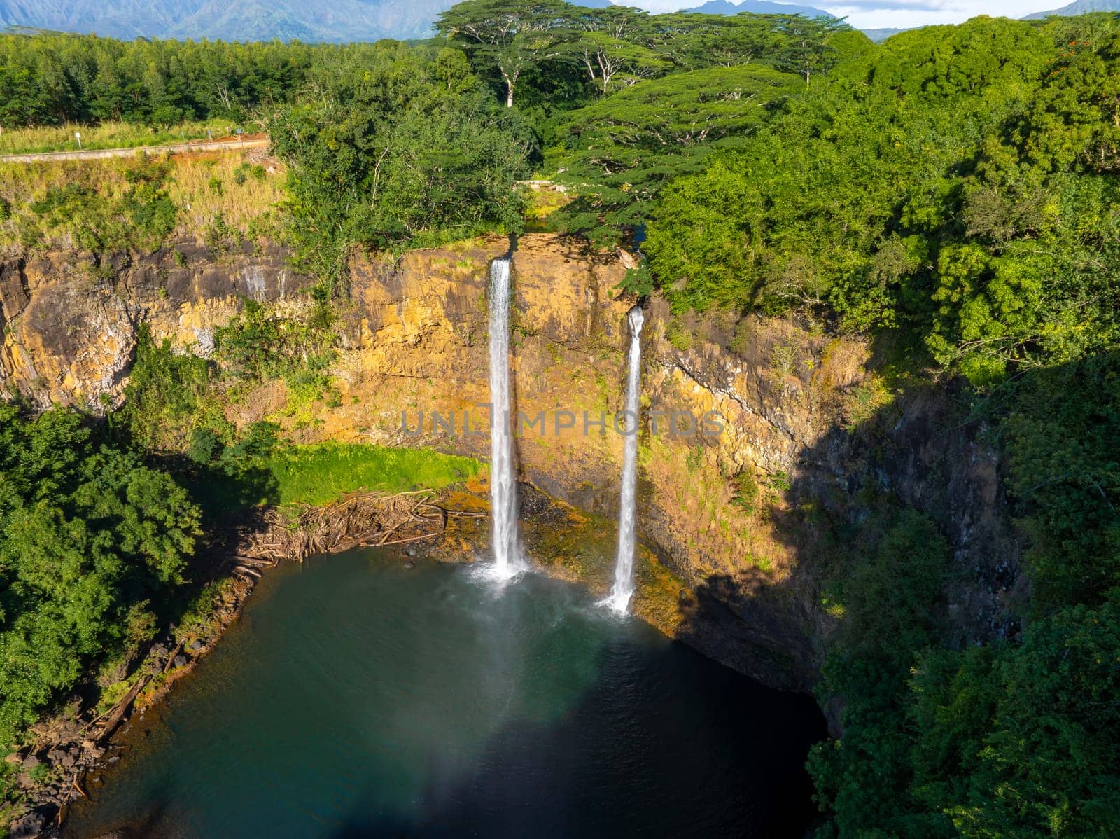 Twin waterfalls of Wailua Falls in Kauai flow into a serene pool, surrounded by lush greenery, rocky cliffs, dense forest, and distant mountains on Kauai.