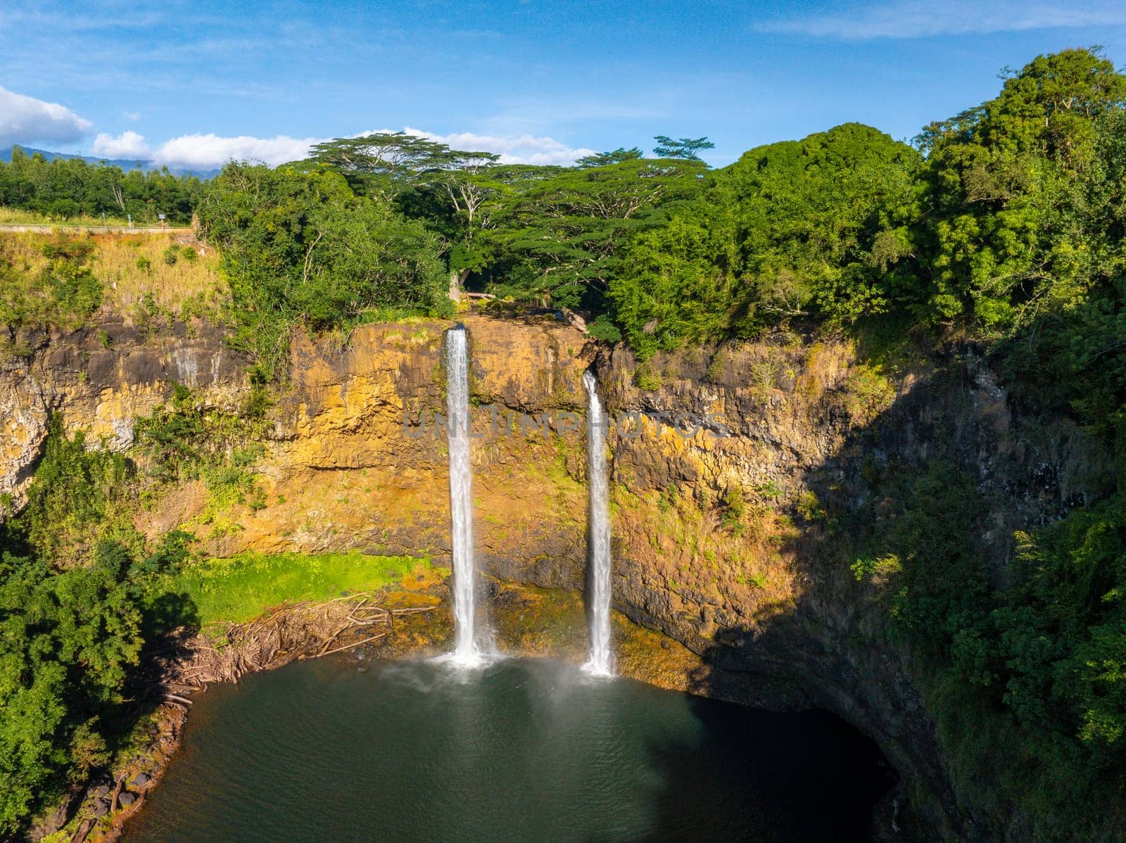 Wailua Falls on Kauai, Hawaii, cascades over a rocky cliff into a circular pool, surrounded by lush greenery under a clear blue sky.