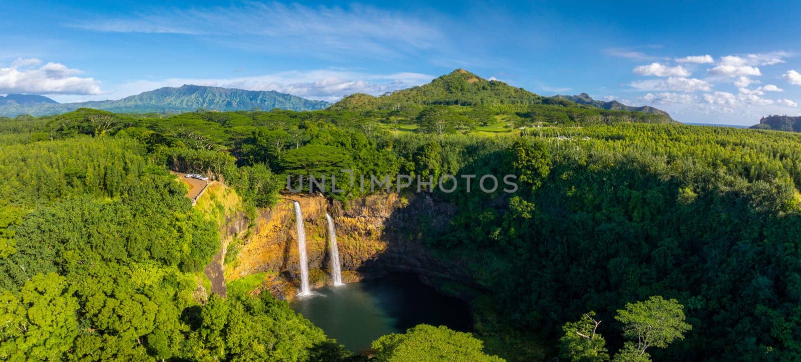 Twin waterfalls cascade into a tranquil pool amid dense tropical foliage, with rolling hills, distant mountains, and a clear blue sky in the background.