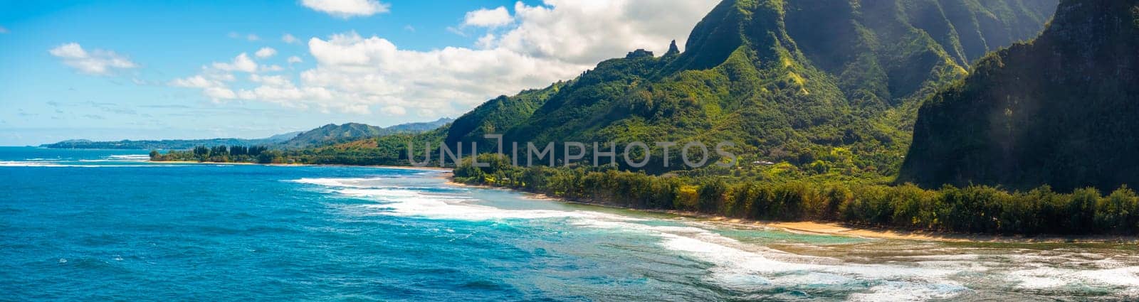 Aerial view of Kauai Island, Hawaii, showing turquoise waters, sandy beach, lush Napali cliffs, and scattered clouds under a vibrant blue sky.