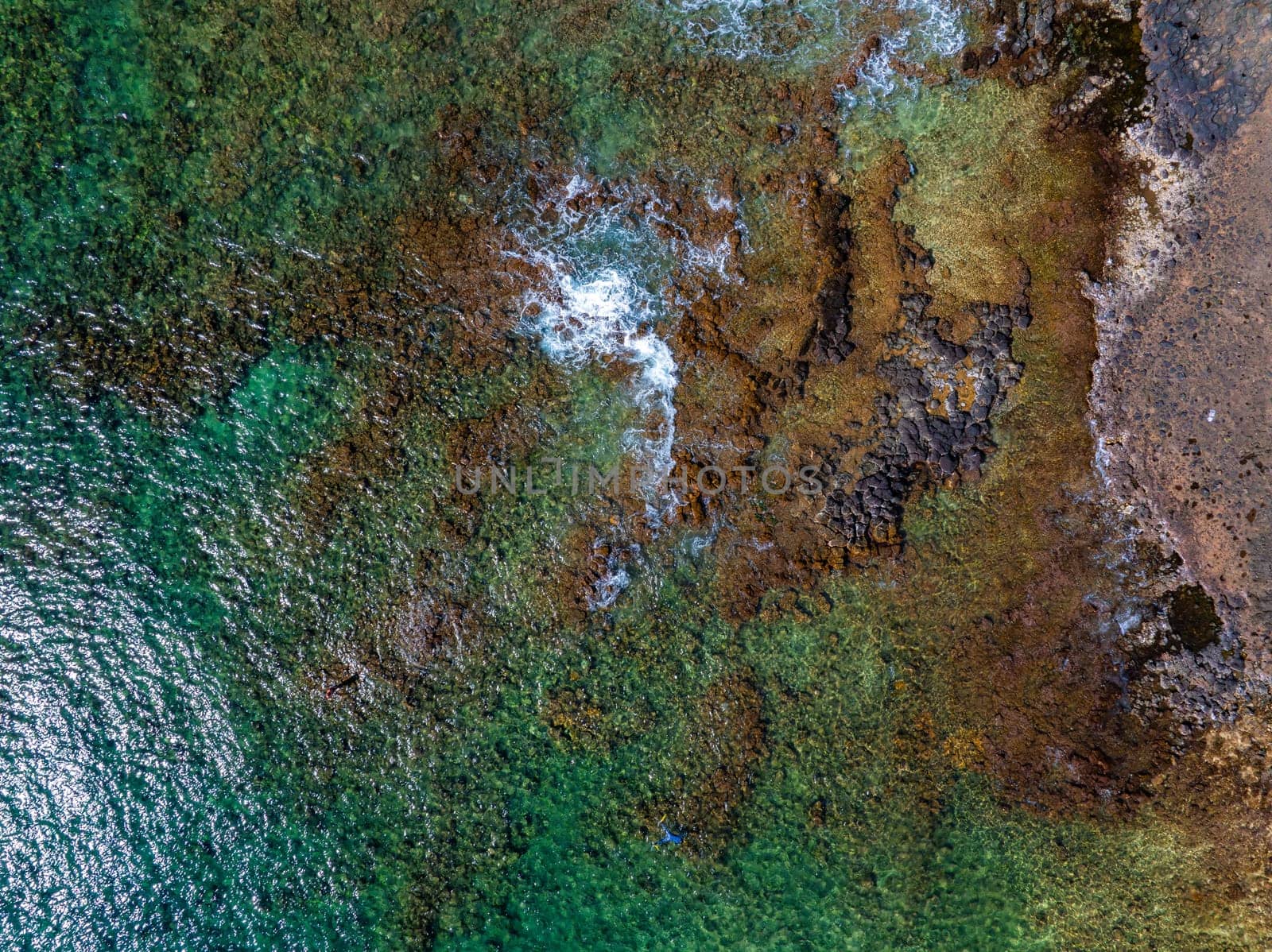 Aerial perspective of Kauai Island's coastline with turquoise waters, volcanic rocks, golden brown sand, and white waves breaking along the shore.