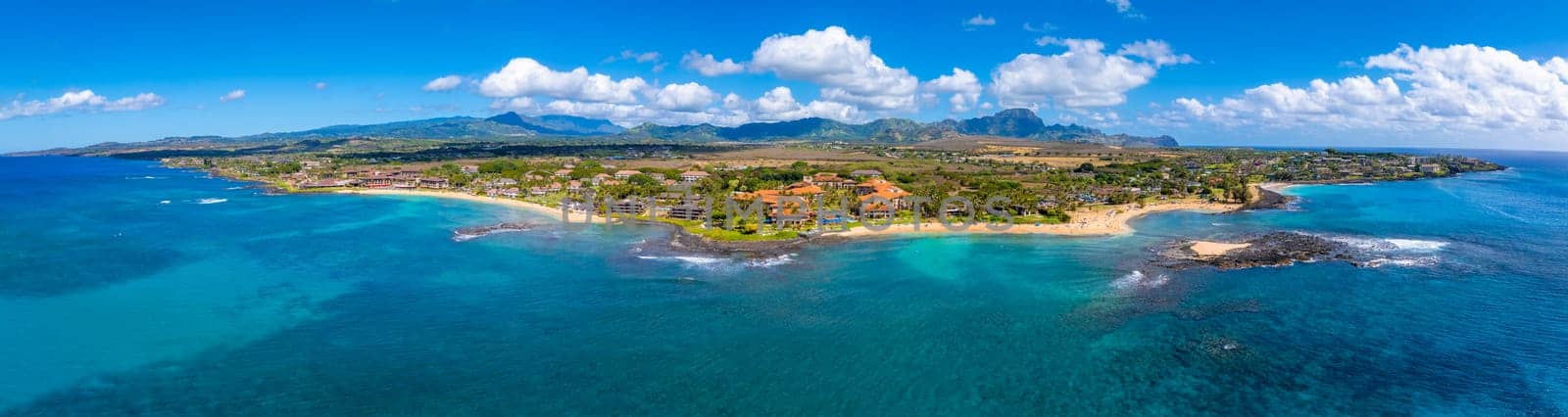 Turquoise ocean meets golden beaches along Kauai's southern coast, with resorts, greenery, rugged mountains, and a bright sky with scattered clouds.